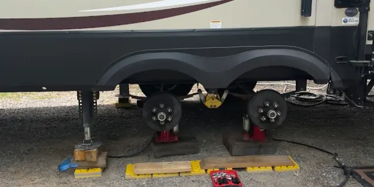 Travel trailer raised on jack stands with both wheels removed, bare hubs exposed, leveling blocks and an impact wrench laid out on the ground