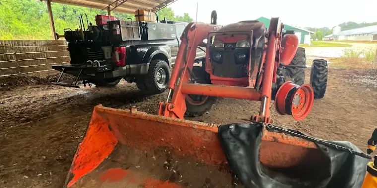 Orange front-loader tractor under a barn with a rear wheel removed and a new tube laid across the loader bucket, Breaux's Roadside service truck parked behind