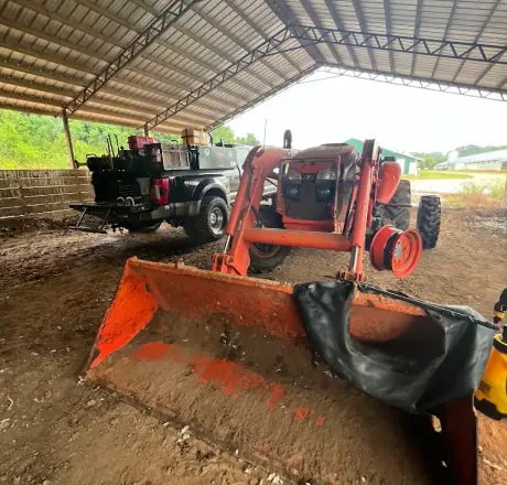 Orange front-loader tractor under a barn with a rear wheel removed and a new tube laid across the loader bucket, Breaux's Roadside service truck parked behind